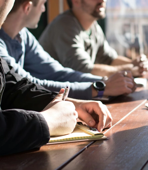 A group of people sitting around a wooden table and one person is writing in a notebook